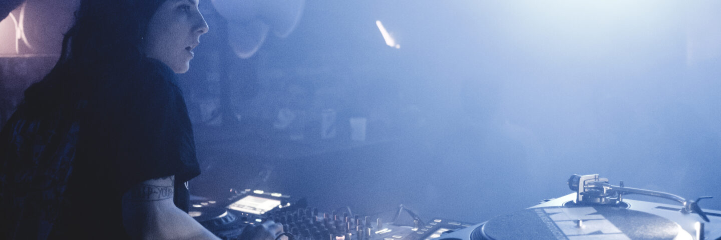 A woman with black hair DJ's in a smokey club.