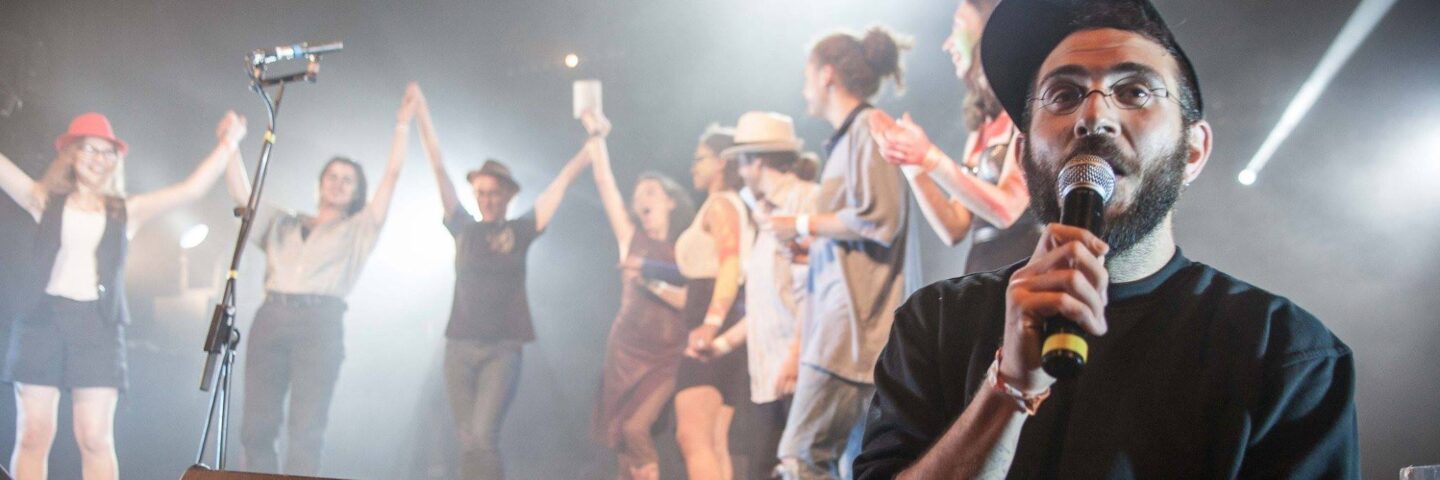 A person sat in front of a choir, in a baseball cap and glasses. They are speaking into a microphone.