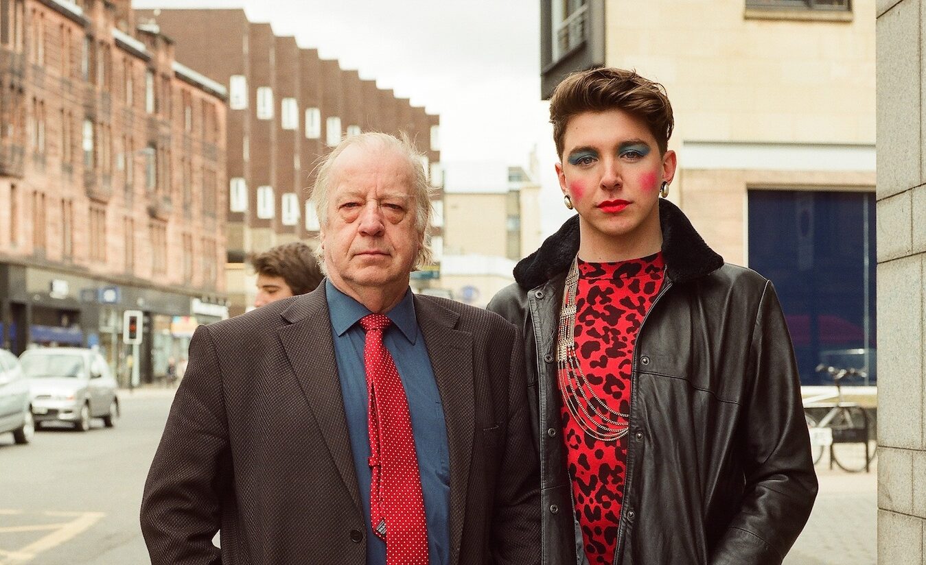 An old man and a young male stands with a male presenting person wearing blue eyeshadow, red lipstick and red blushed cheeks.