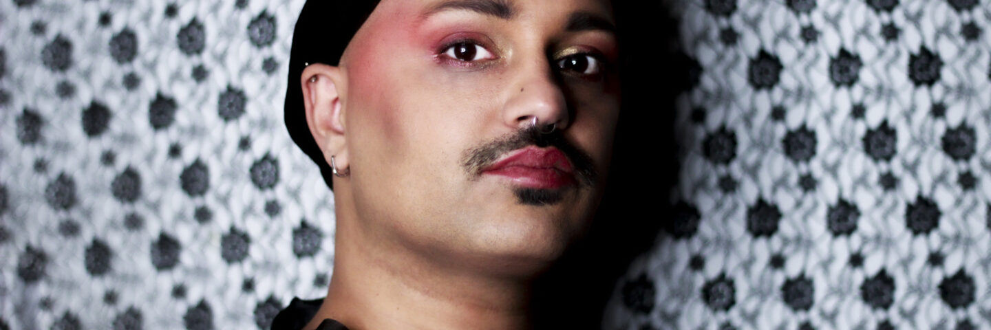 A British Asian person with a moustache in pink eye makeup and red lipstick, on a black and white patterned background. 