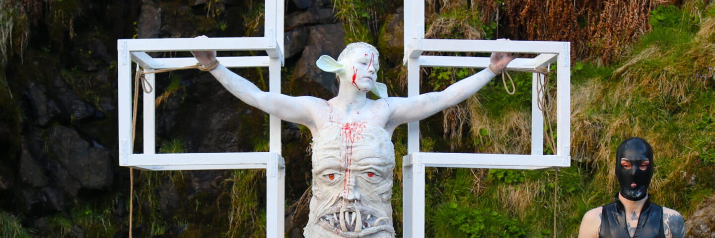 A person in white prosthetics and white body paint is tied inside a white cross. They are seen in a leafy landscape, next to a person in black latex. 