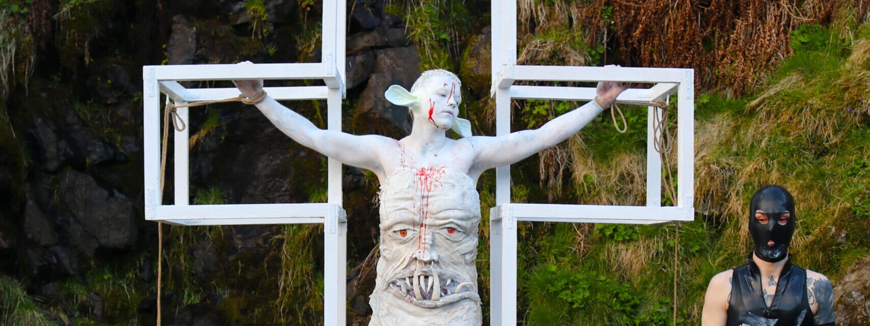 A person in white prosthetics and white body paint is tied inside a white cross. They are seen in a leafy landscape, next to a person in black latex. 
