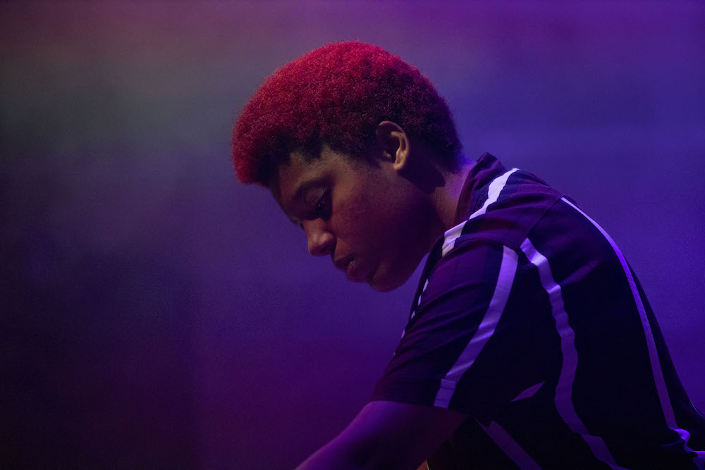 A black woman who is side on to the camera looking down at her music equipment. The picture is lit with a purple light