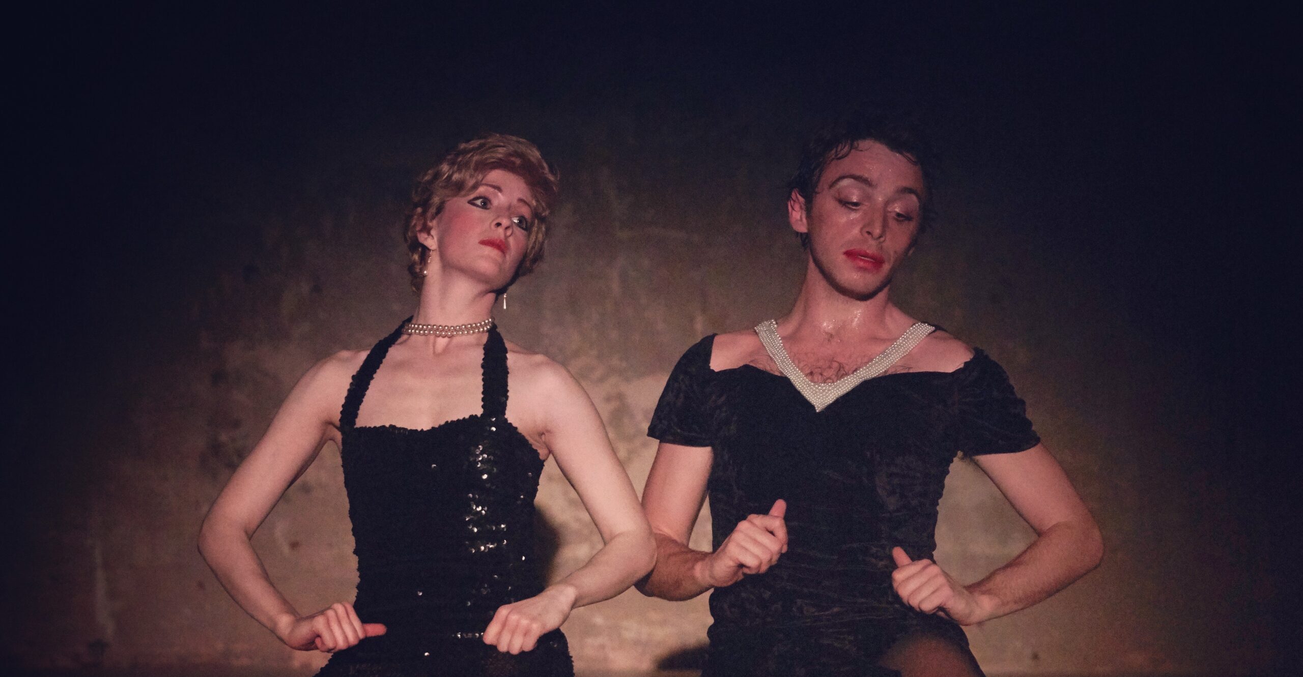 Two white performers sit on a wooden bench in black formal dresses and high heels. Daniel wears red lipstick and a necklace. Eleanor wears a blond wig like Princess Diana.  