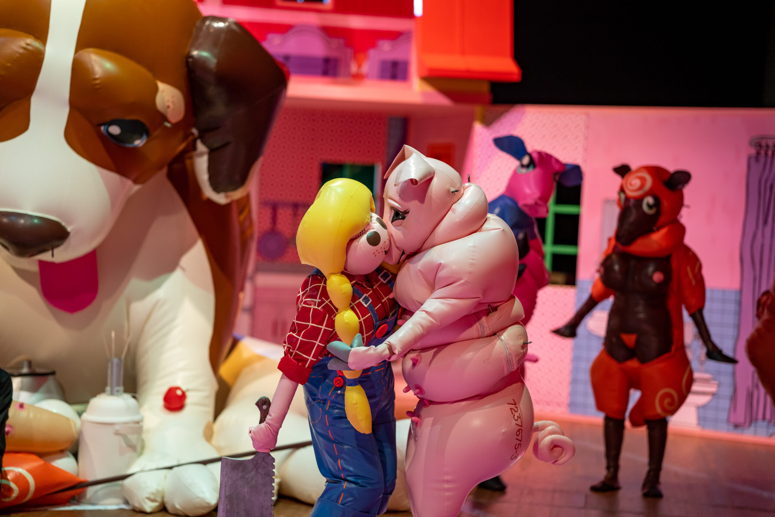 A person in a brightly coloured latex farm girl outfit embraces a person in a pink latex pig outfit. 