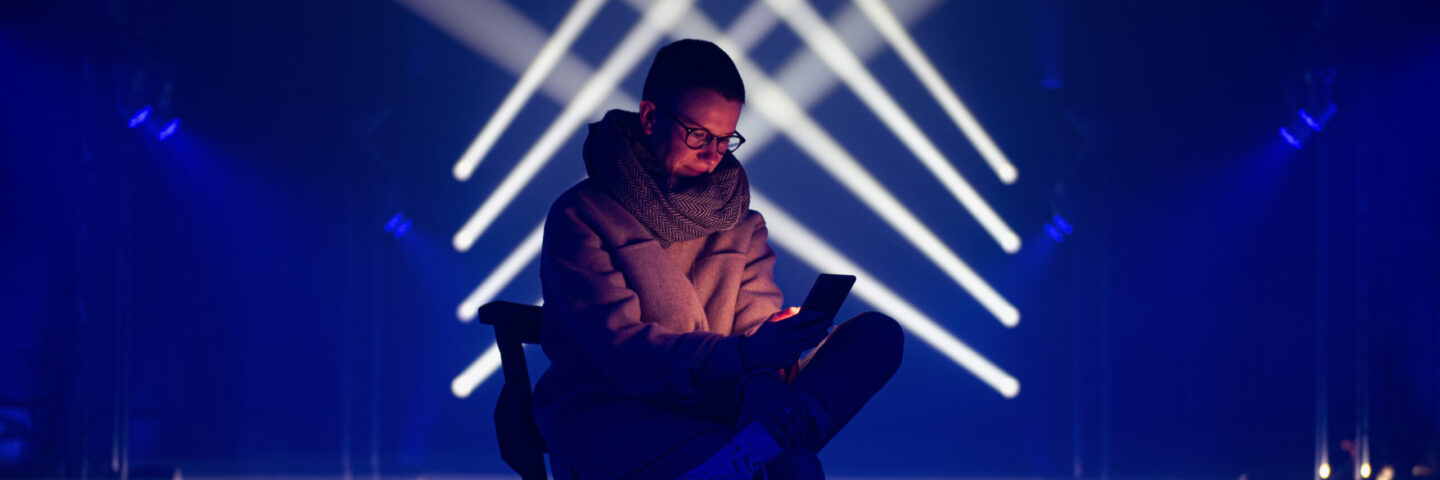 A woman with cropped hair and glasses sits within beams of blue and white light. 