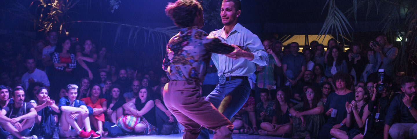 Two men dancing in pink and purple light. One is facing the camera wearing a blue shirt. The other has his back to the camera and slicked back brown hair, and a floral short.