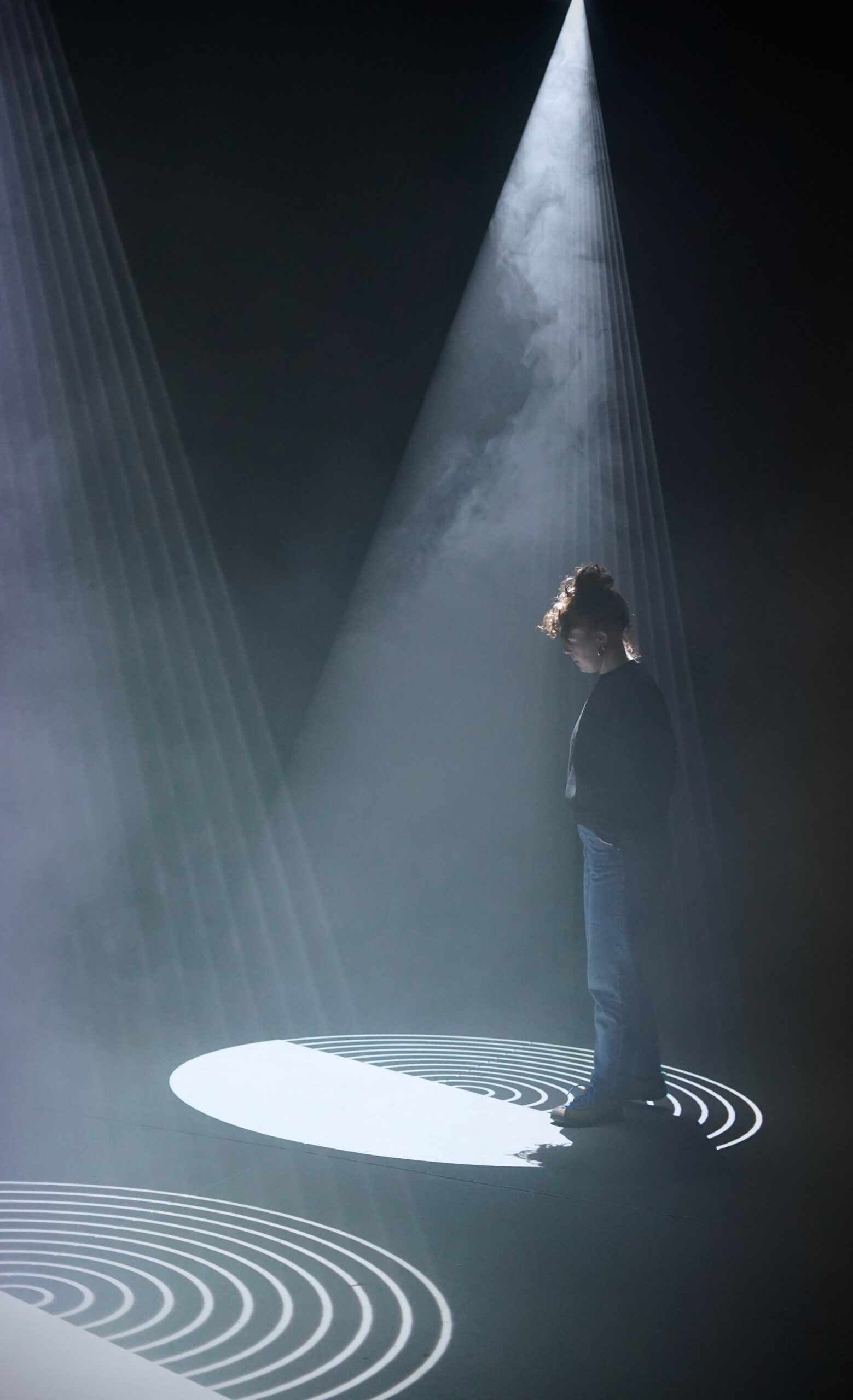 A white woman in a black jumper and blue jeans stands in a pool of white light and haze.  