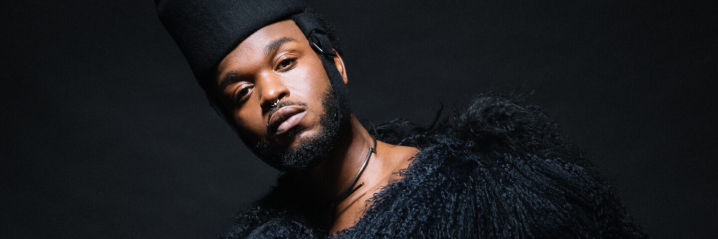 Young black artist Cakes Da Killa wears a gorgeous black fur and fringe outfit and hat. He looks directly at the camera, posing on a black background. 