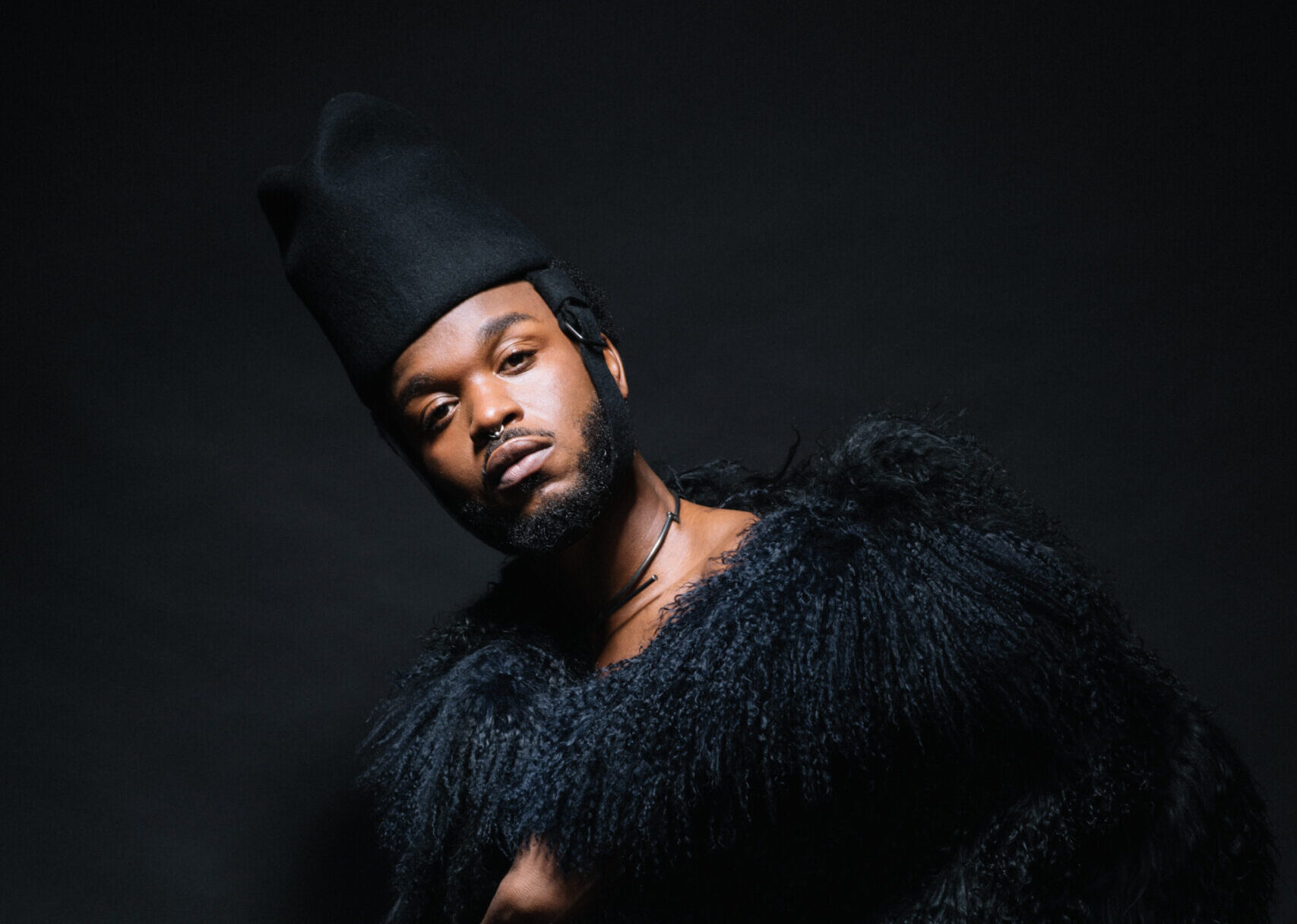 Young black artist Cakes Da Killa wears a gorgeous black fur and fringe outfit and hat. He looks directly at the camera, posing on a black background. 
