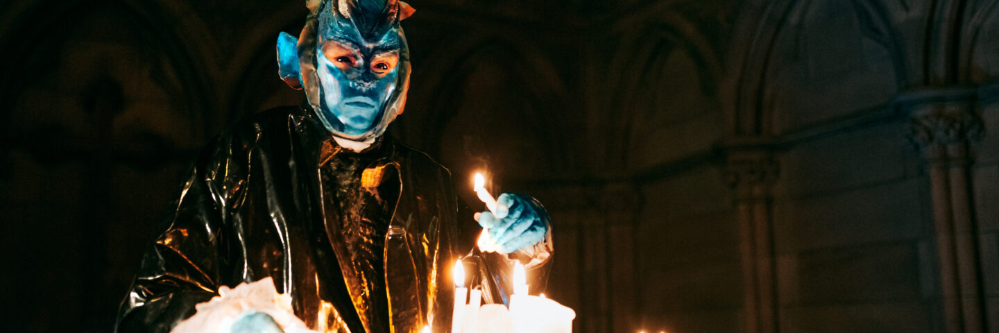 An orge character stands above a number of lit candles, in a church