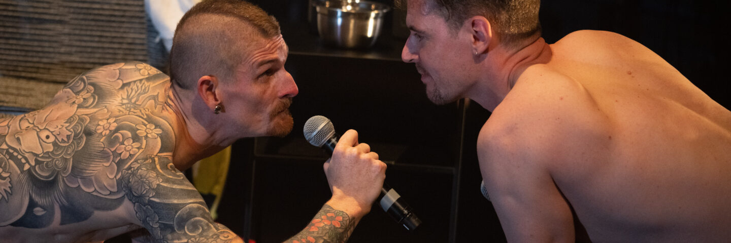 A white man with a mohawk and tattoos is on all fours holding a microphone. He faces another white man with brown hair. They are both shirtless.  