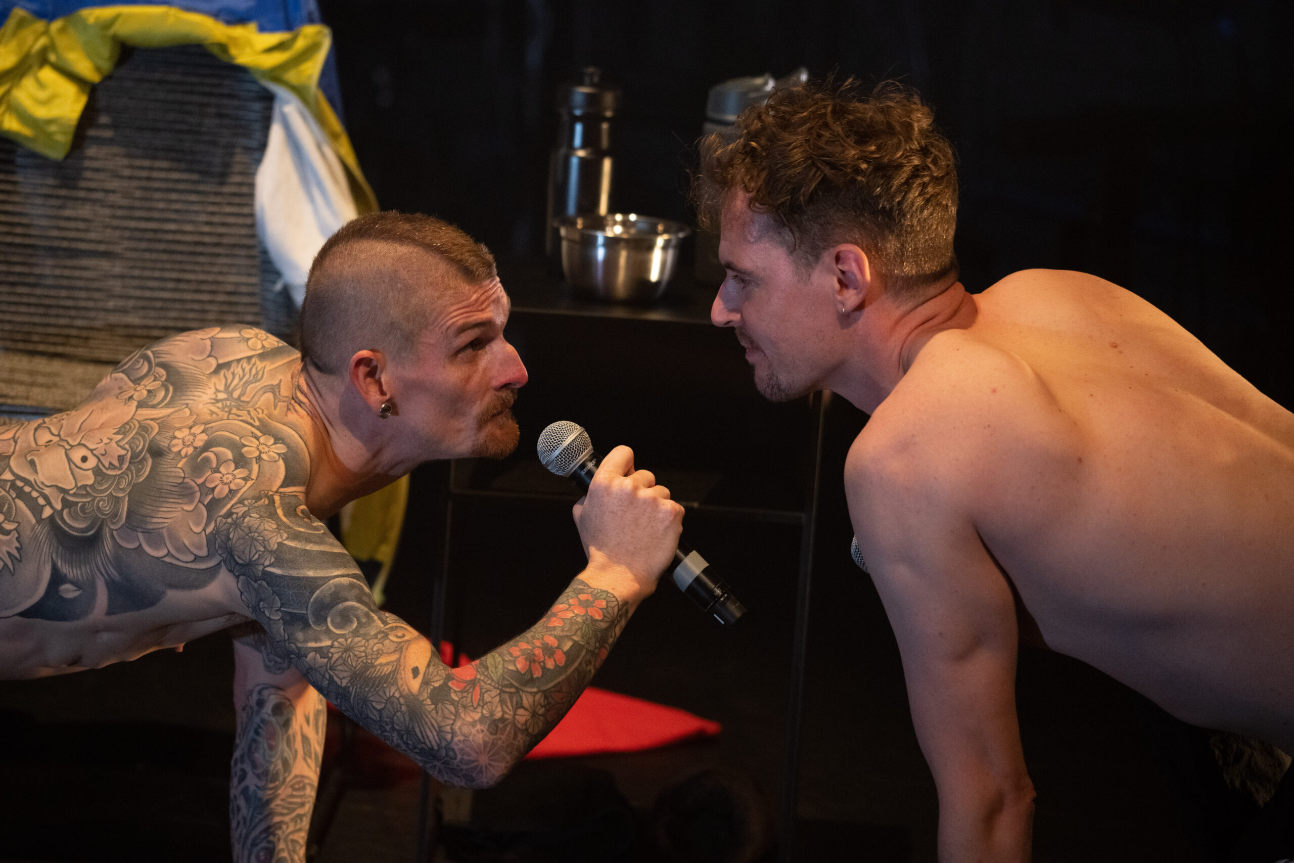 A white man with a mohawk and tattoos is on all fours holding a microphone. He faces another white man with brown hair. They are both shirtless.  