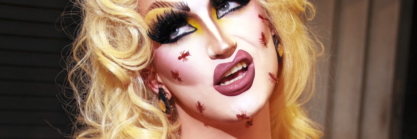 A white drag queen wears a blond wig and a cockroach inspired outfit. She has cockroaches on her face.