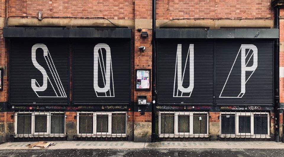 Four black shutters on a red brick building. On each shutter in white text is a letter of the word soup.