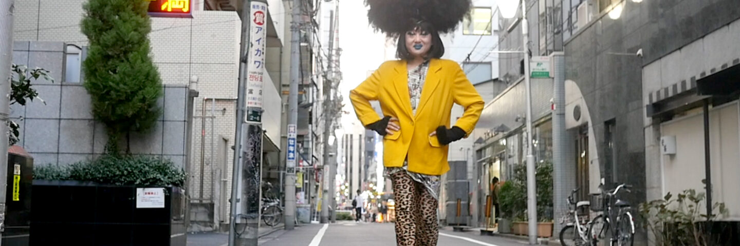 A drag queen stands in the streets of Tokyo, wearing big black hair and a yellow jacket. 