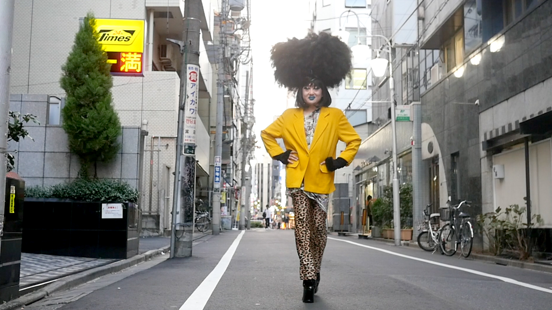 A drag queen stands in the streets of Tokyo, wearing big black hair and a yellow jacket. 