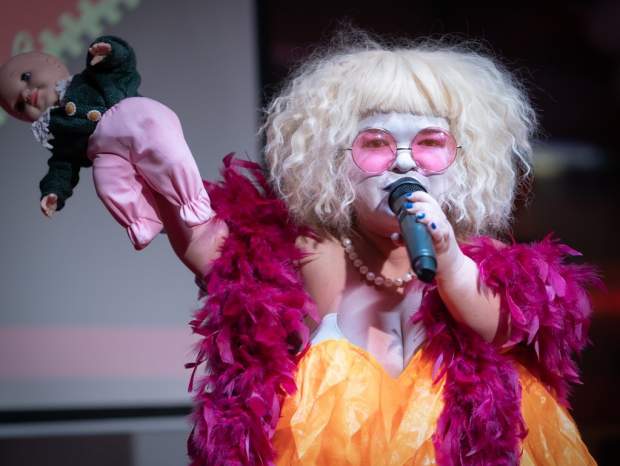 The artist Tammy Reynolds wears a blond wig, orange dress, red feather bower and pink sunglasses. They are holding a microphone and a children's doll.