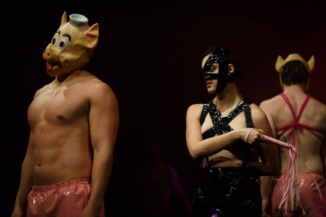 A white person in black latex clothing holds a flogger near two white people in pink fetish clothing and pig masks. 