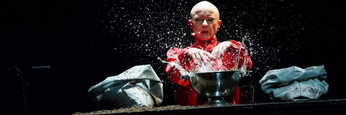 A bald white woman sits with a table in front of her. The table has 2 blue bags on it in at either end and a silver bowl in the centre. The woman's hands are splashing water from the bowl.