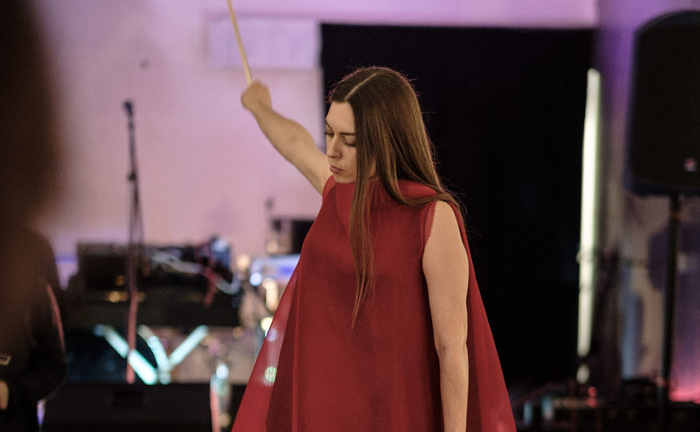 A white woman in lond red dress stands on a stool. She holds a violin. One hand is in the air.