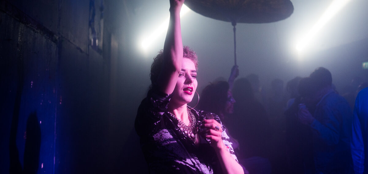 A white person in a nightclub holds a decorated parasol above their head. 