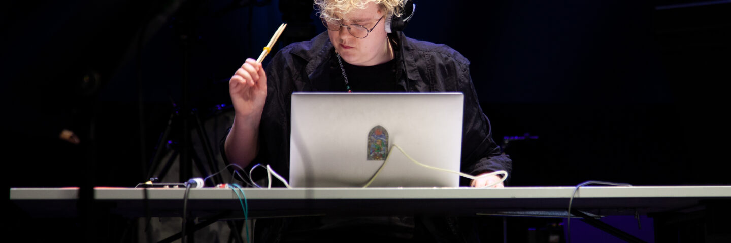 A person sits at a desk with a silver laptop. They have blond wavy short hair and black headphones on. There are wires lying on 