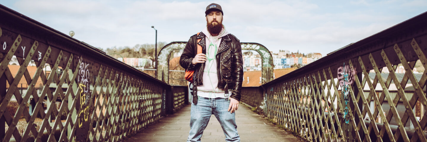 A white man in blue jeans, a pink hoodie and a leather jacket stands on a bridge. He has a big beard. 