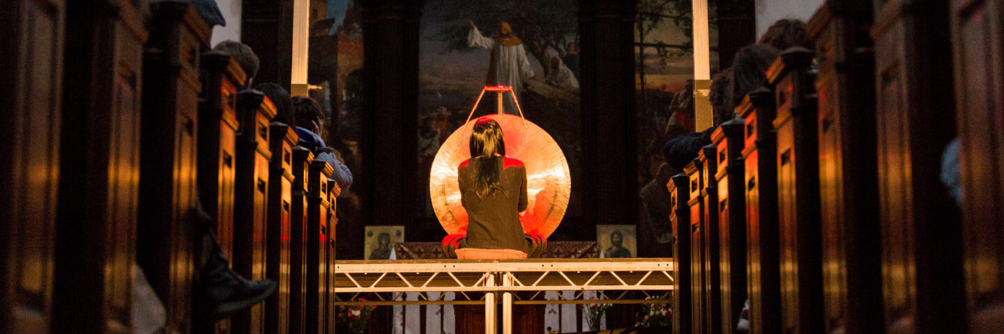 The artist Rrose sits infront of a large metal gong, in a beautiful church. She wears a black wig and black dress. Her back is turned. 