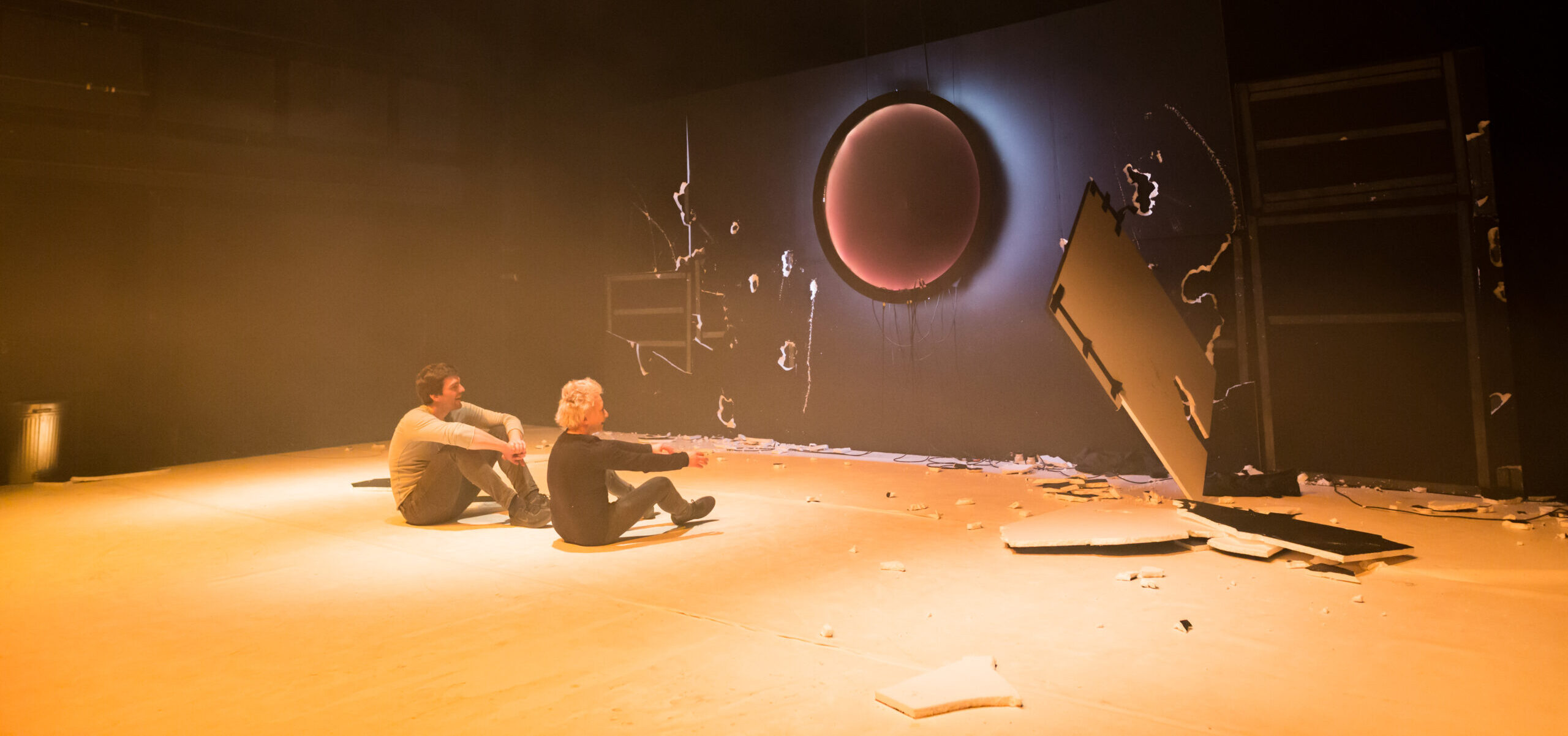 Two white people sit looking at a destroyed black plasterboard wall as part of a performance. 
