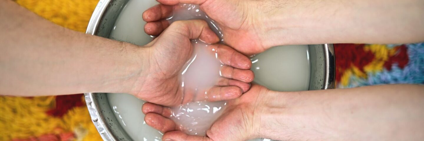 Two pairs of white hands are seen in a bowl of white fluid. The fluid is in a metal bowl, on a yellow rug. 