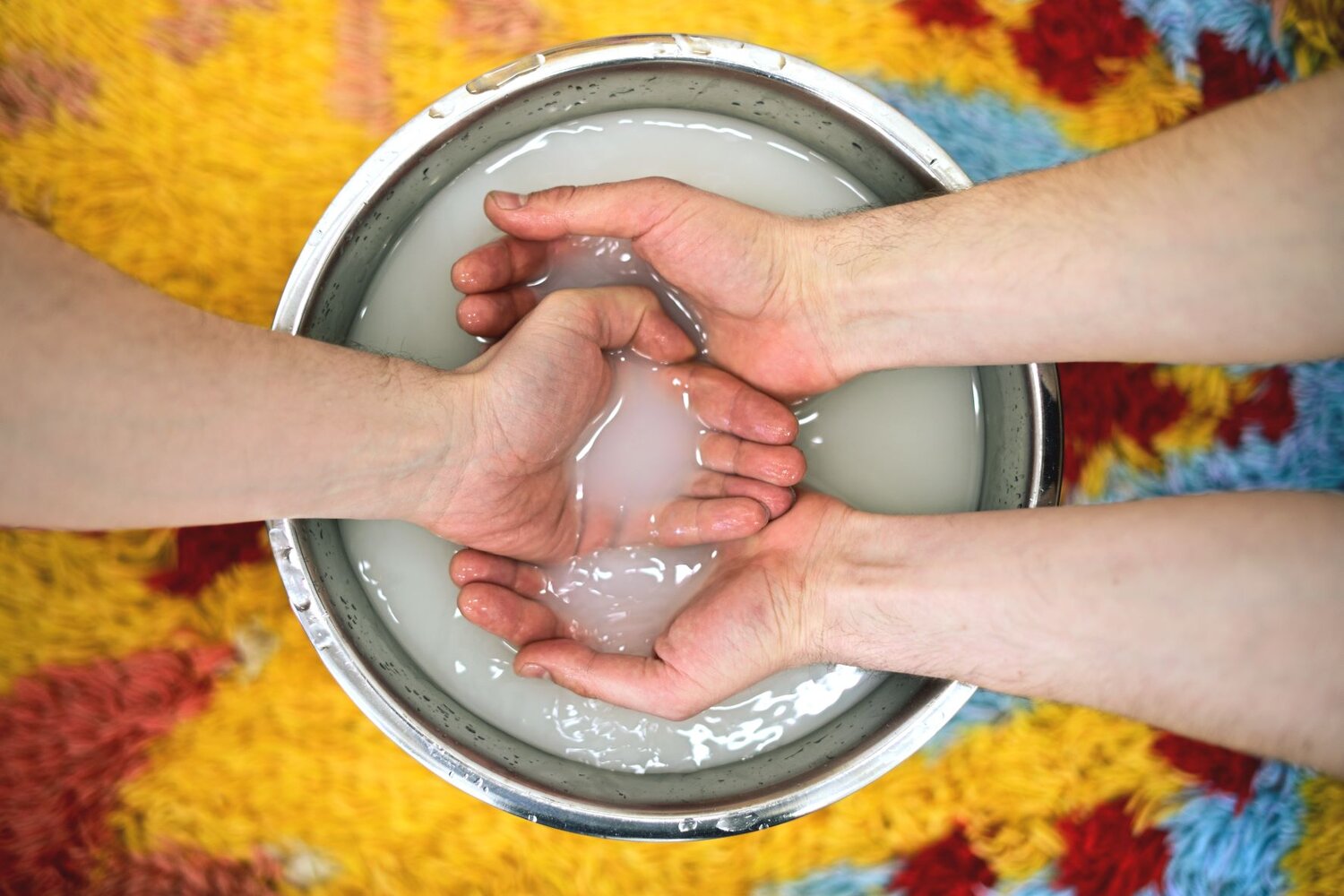 Two pairs of white hands are seen in a bowl of white fluid. The fluid is in a metal bowl, on a yellow rug. 
