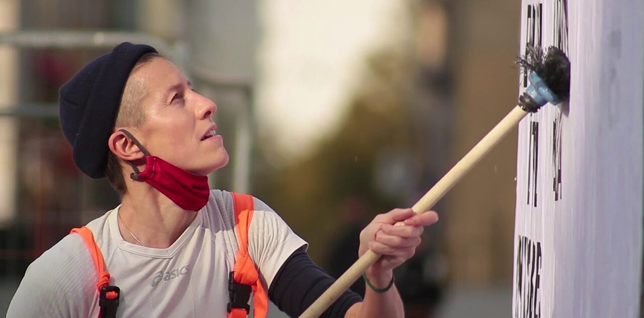 A white woman in hi-vis and a beenie hat pastes a poster to a hoarding. 