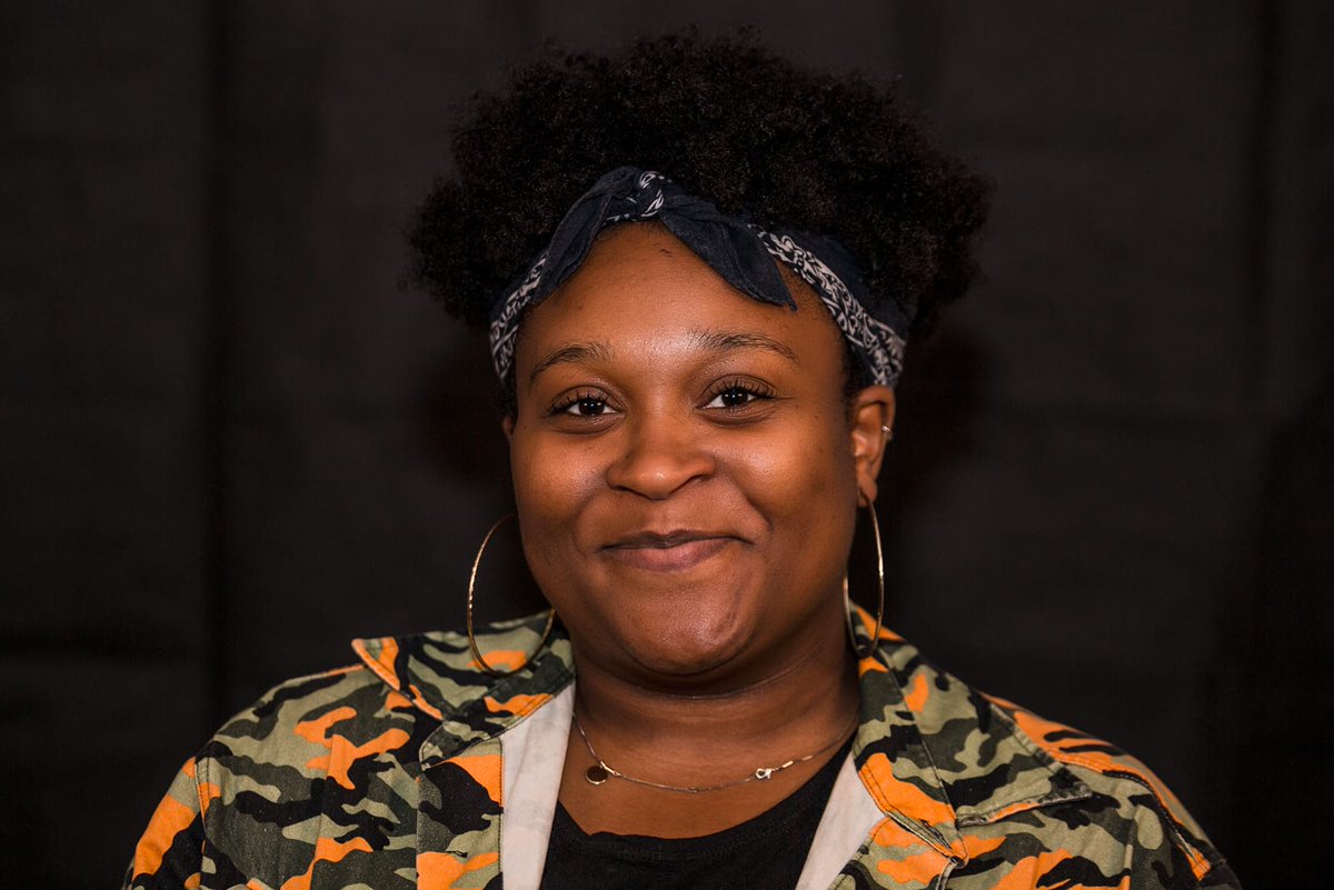 A black woman wears a neon camo print top, a necklace and a headscarf. She is smiling at the camera 