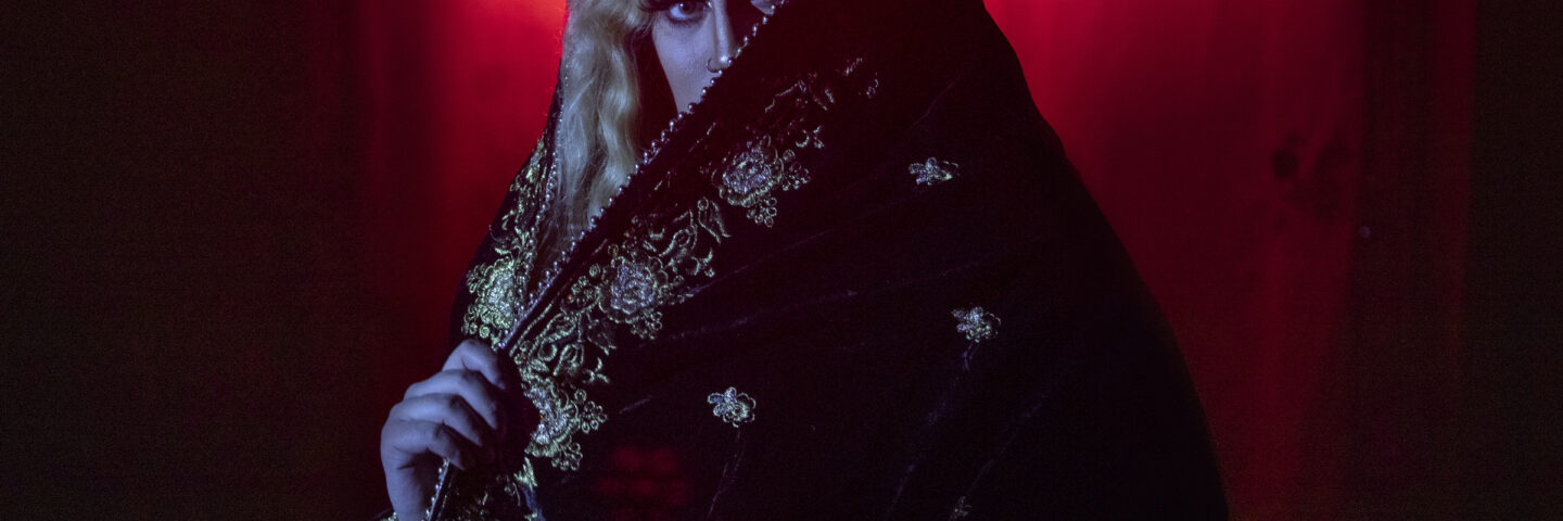 South Asian drag queen Val Qaeda peers from behind an ornate cloth. She wears a blond wig, and is lit in red and white.  