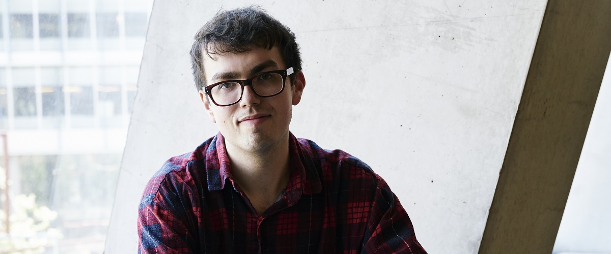 A white man with short brown hair and black square glasses stands face on to the camera. They wear a dark red and blue plaid shirt. The background behind them is grey concrete. 