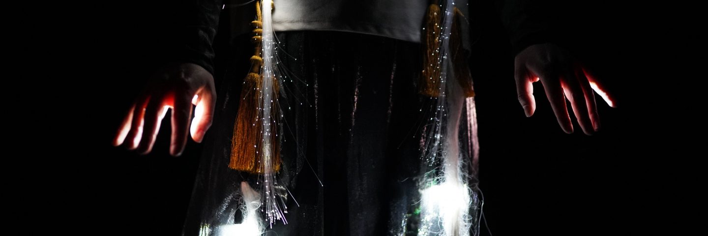 A person's middle torso is seen in darkness. They have a white top and long braids. Fibre-optic lights are entwined into their braids and light pours from their fingers. 