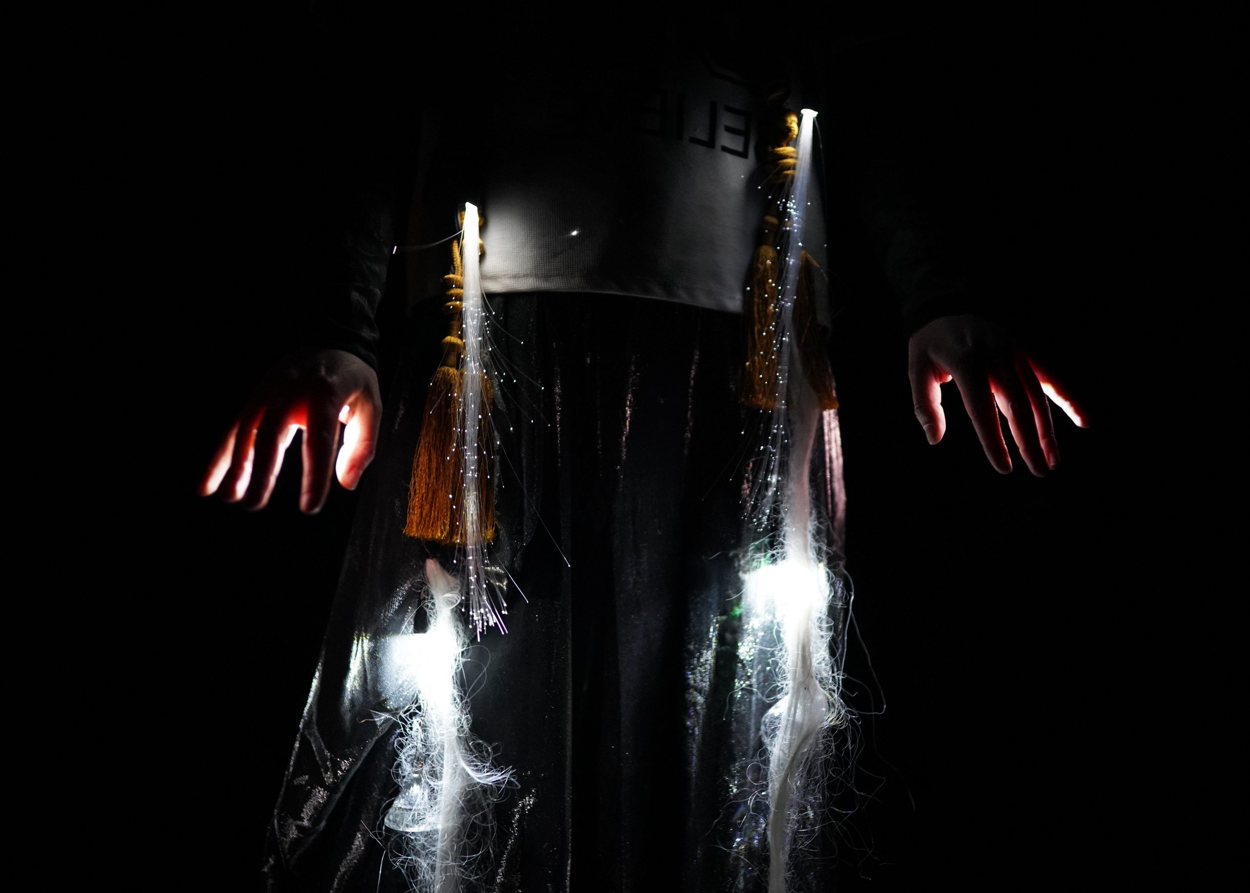 A person's middle torso is seen in darkness. They have a white top and long braids. Fibre-optic lights are entwined into their braids and light pours from their fingers. 