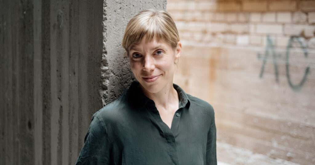 A white woman with brown hair and a green shirt stands against a brick wall. She is looking at the camera.