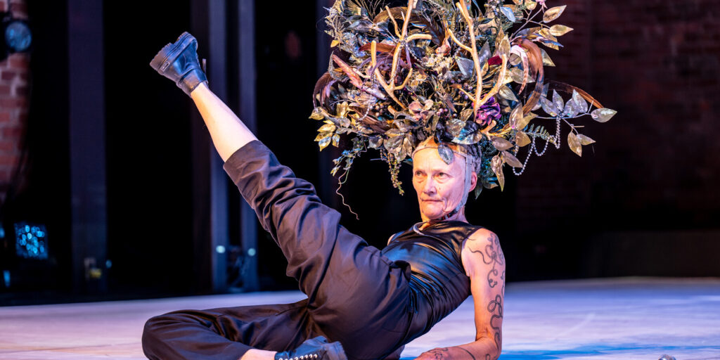 An older white women is seen laying on a stage. She kicks her lick in the air, and wears an extravagant headpiece which looks like the brances of trees.