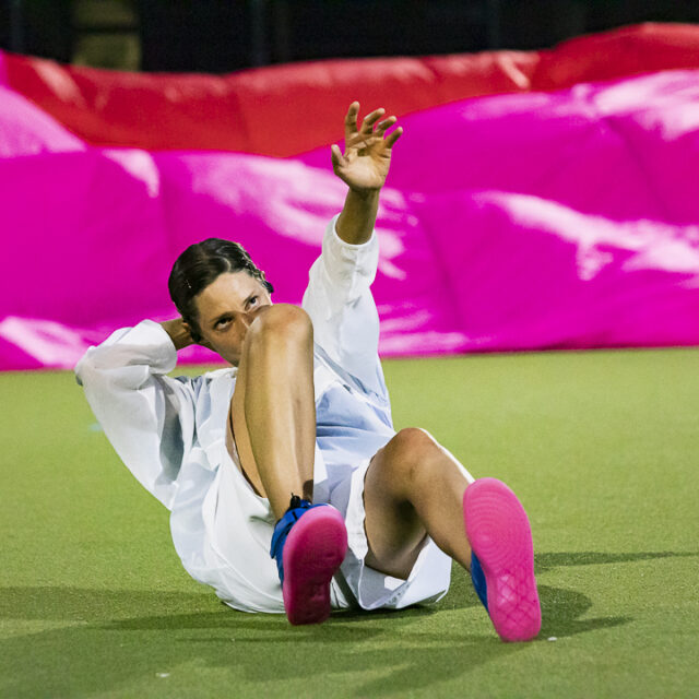 A woman with brown hair, white tracksuit and pink shoes is seen on a football pitch, with pink parachute material behind her.