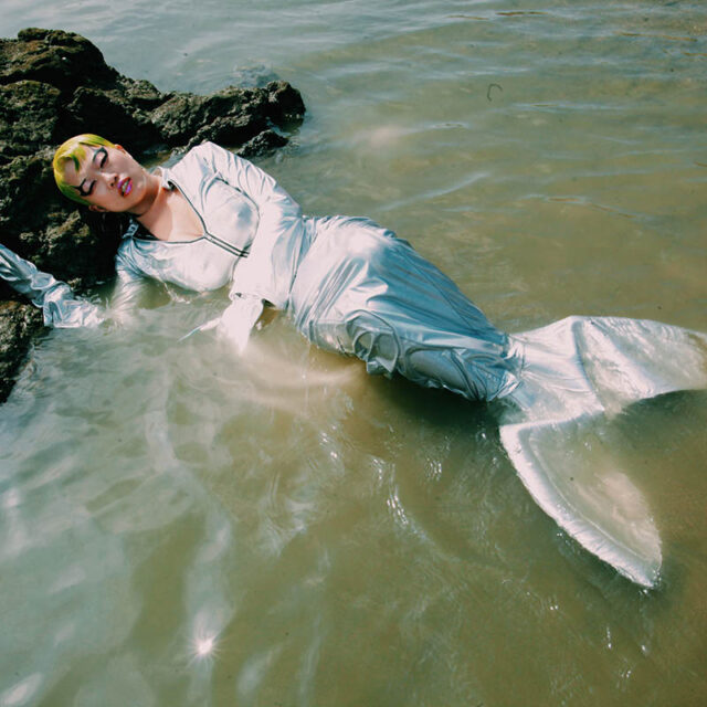 An Asian woman with yellow hair and a blue and silver mermaid tail is pictured in the sea, with her head on a rock.
