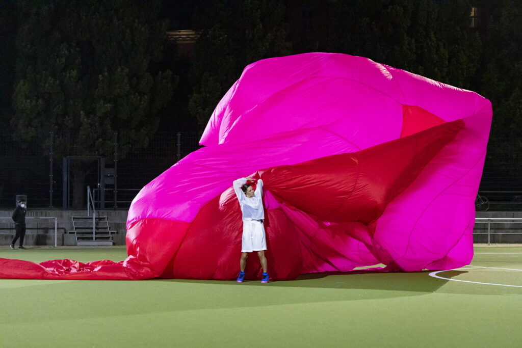 The artist Milla Koistinen is a white woman wearing a white sports outfit. She stands on a football pitch, manipulating a giant pink parachute sculpture.