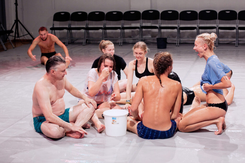 A group of people sit in swimwear on a dancefloor covered in lube.